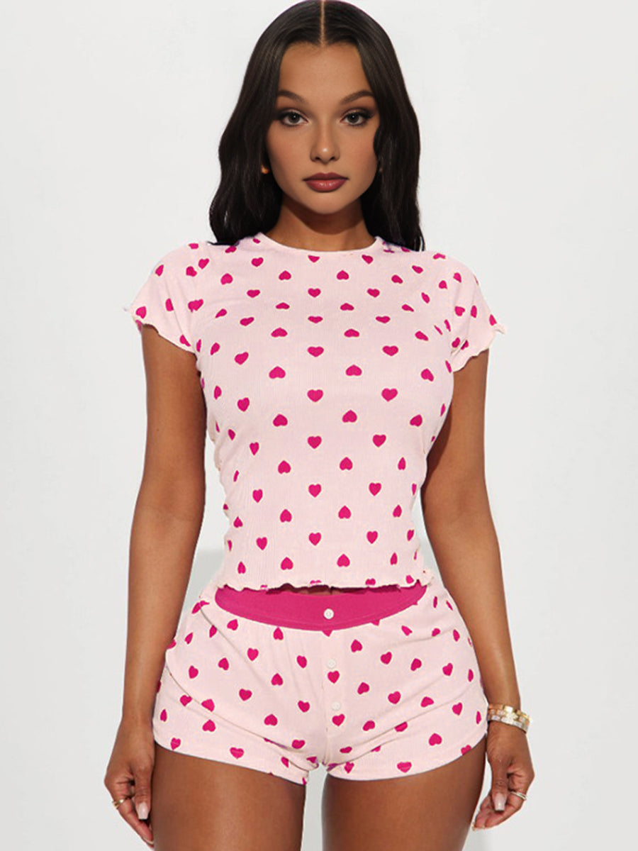 A woman wearing the Costume Inspire All My Love heart print pajamas set, featuring a pink rib-knit short sleeve crop top and matching high-waisted shorts with a fuchsia waistband.